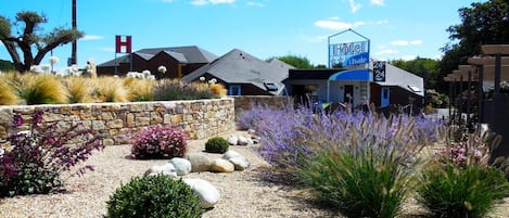Hotel con un letrero azul, pared de piedra y camino de grava con varias plantas.