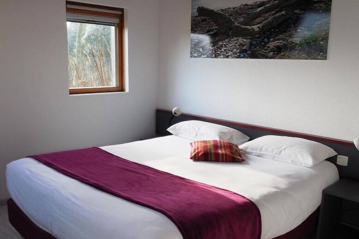 A bedroom with a large bed, a window, a painting, and a nightstand.