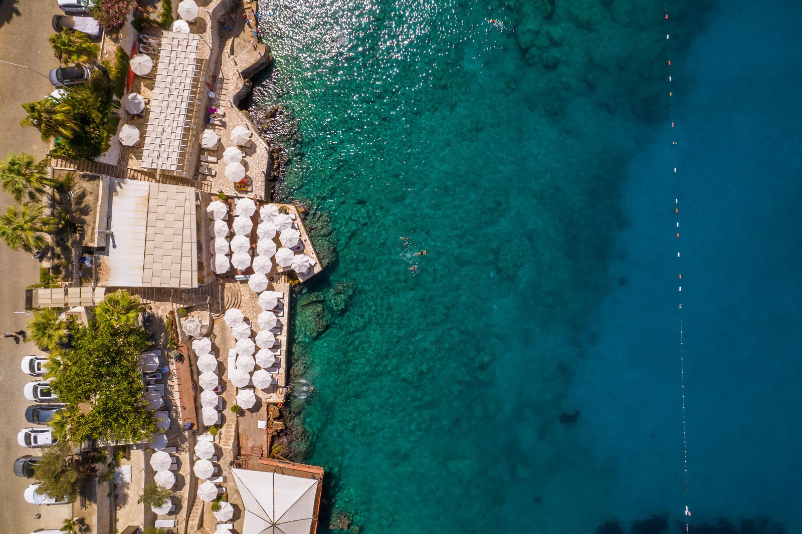 An aerial view of a beachside resort with rows of sun loungers and umbrellas, a paved walkway, and a clear blue sea.
