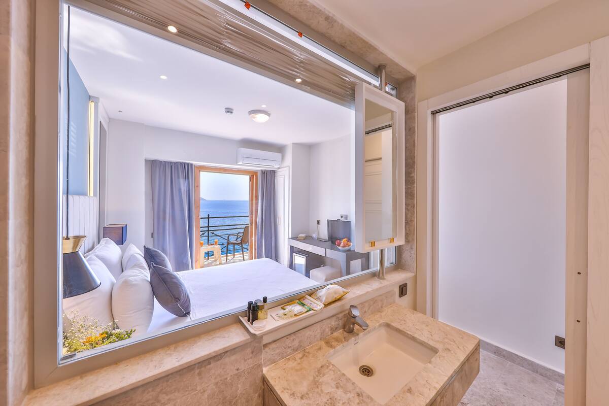 Una habitación de hotel moderna con una cama grande, un baño con lavabo y espejo, y un balcón con vista al mar.