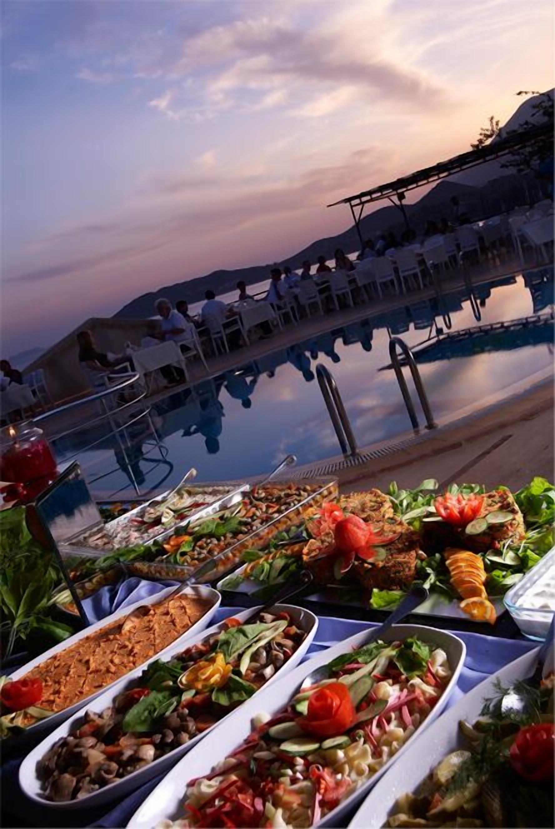 A buffet table with various dishes, a poolside seating area, and a view of the mountains in the background.