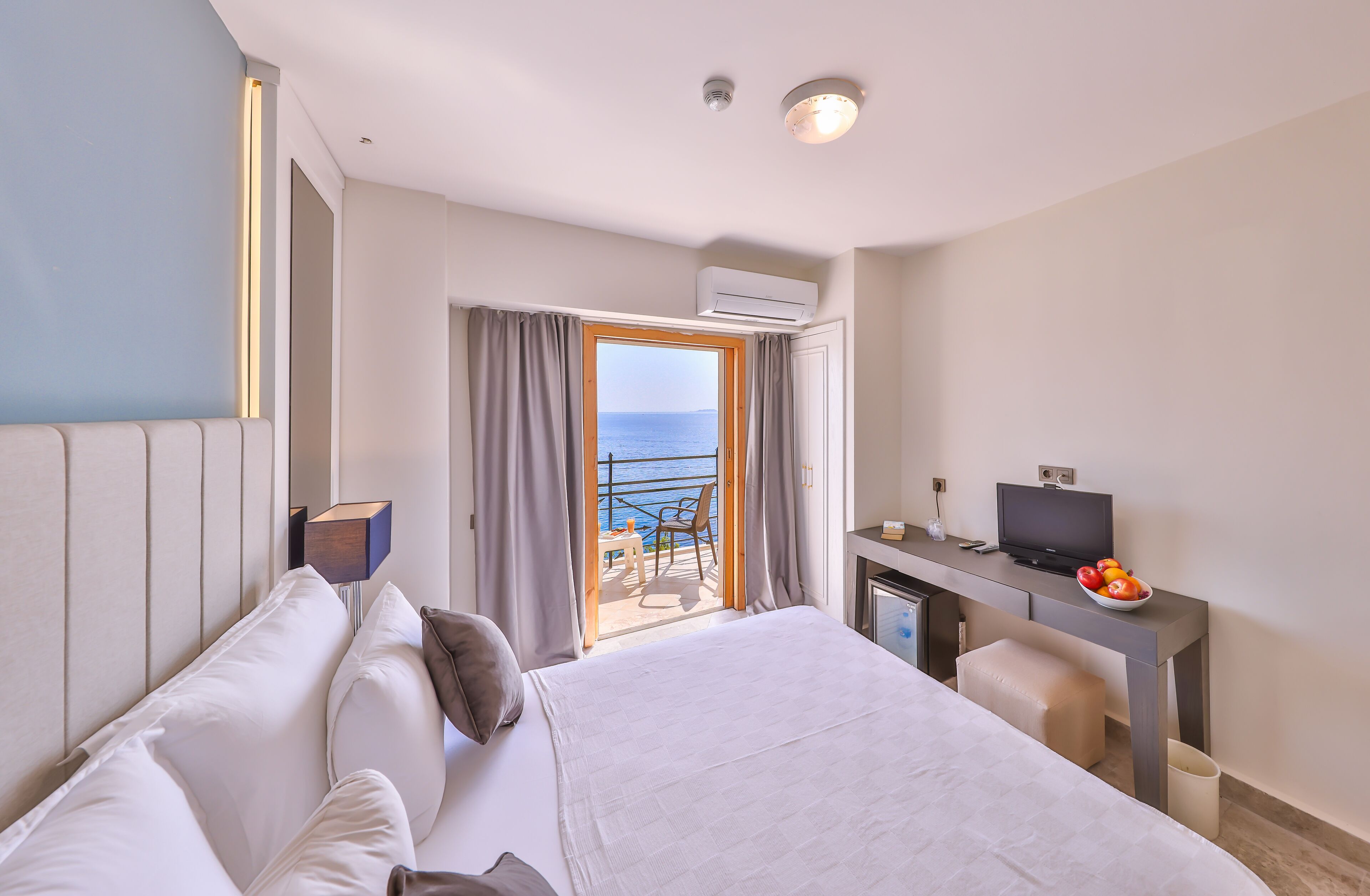 Een hotelkamer met een bed, een bureau met een televisie en een balkon met uitzicht op zee.