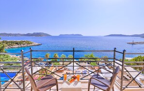 Un balcon avec vue sur la mer, des chaises longues, une table avec un assortiment de fruits et des boissons, et une piscine en arrière-plan.