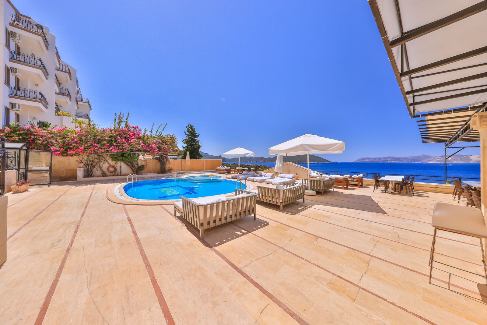 A pool area with lounge chairs and umbrellas, a clear view of the sea, and a building with balconies.