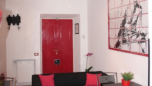 Une pièce avec une porte rouge, un canapé noir orné de coussins rouges, une petite table sur laquelle est posée une plante en pot, et un mur décoré d’une grande œuvre d’art en noir et blanc.