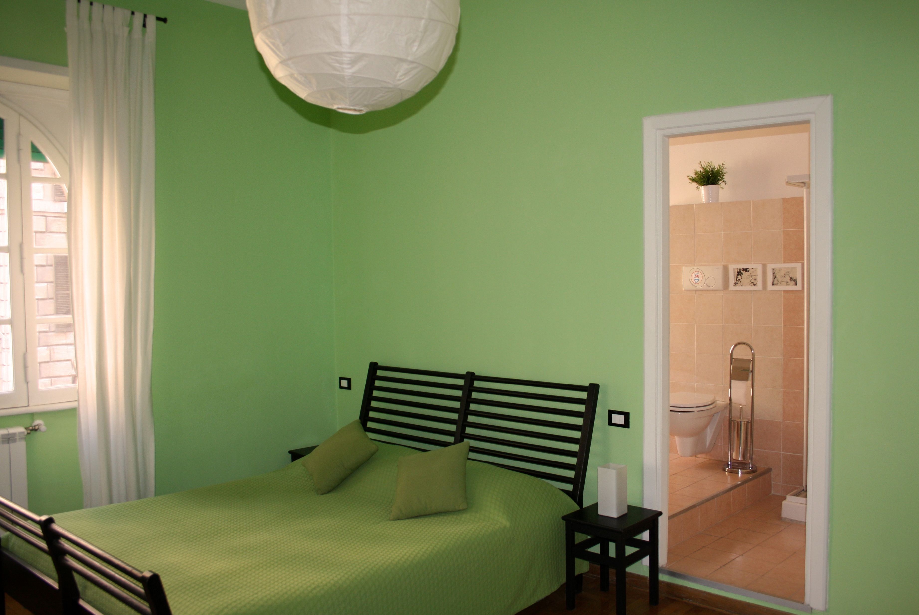 Een slaapkamer met een bed, nachtkastje, raam met gordijnen en uitzicht op een badkamer.