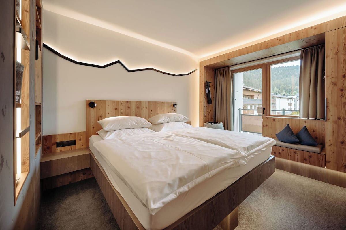 A bedroom with a wooden headboard, a large bed, a window with curtains, and a view of buildings outside.