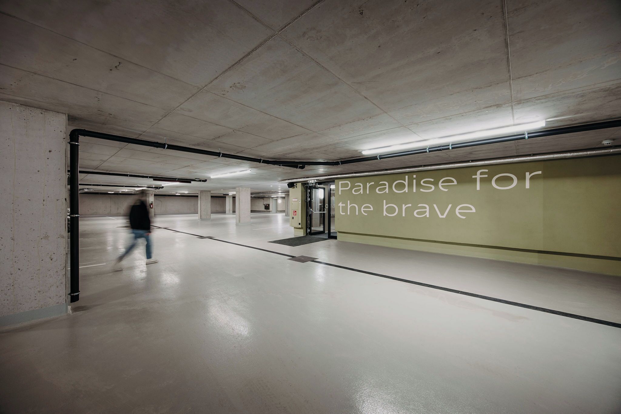 Un vaste espace de stationnement souterrain, bien éclairé, doté d’un sol en béton propre et d’un mur sur lequel est inscrit le texte : « Paradis pour les courageux. »