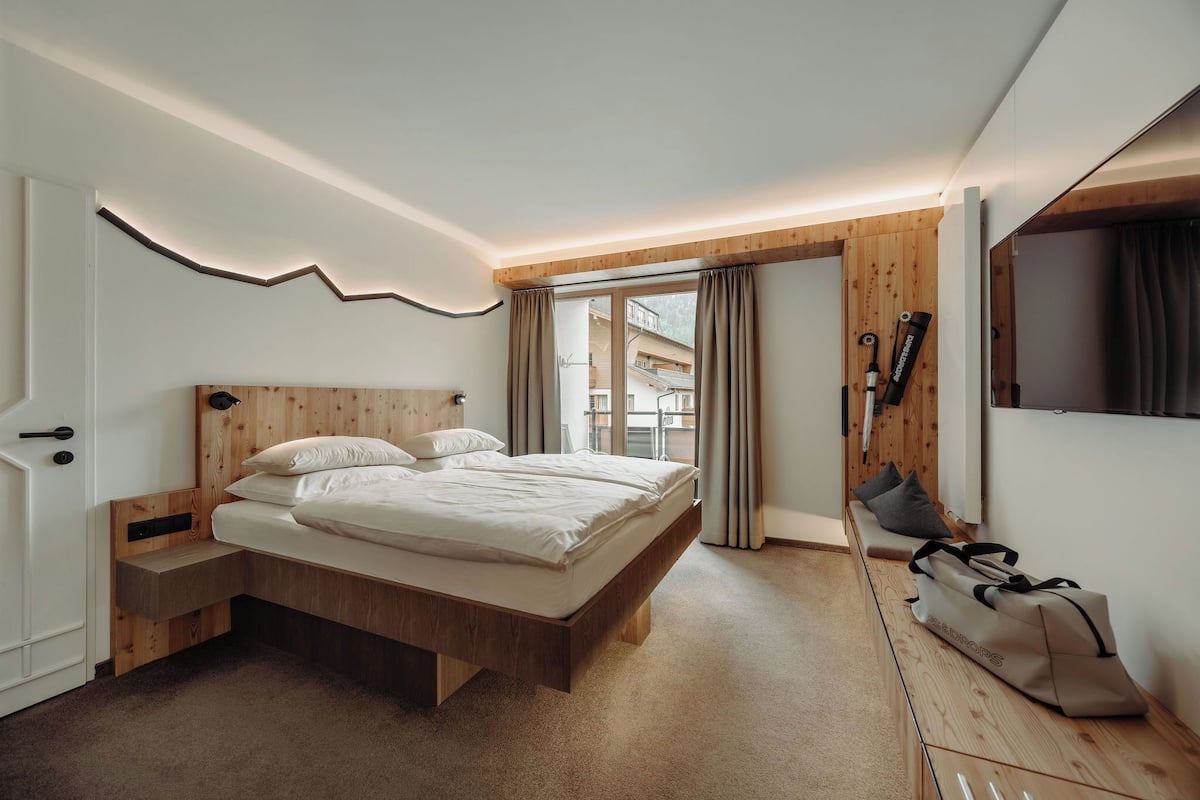 A modern bedroom with a wooden bed, a built-in bench, a suitcase, and a view of a balcony.
