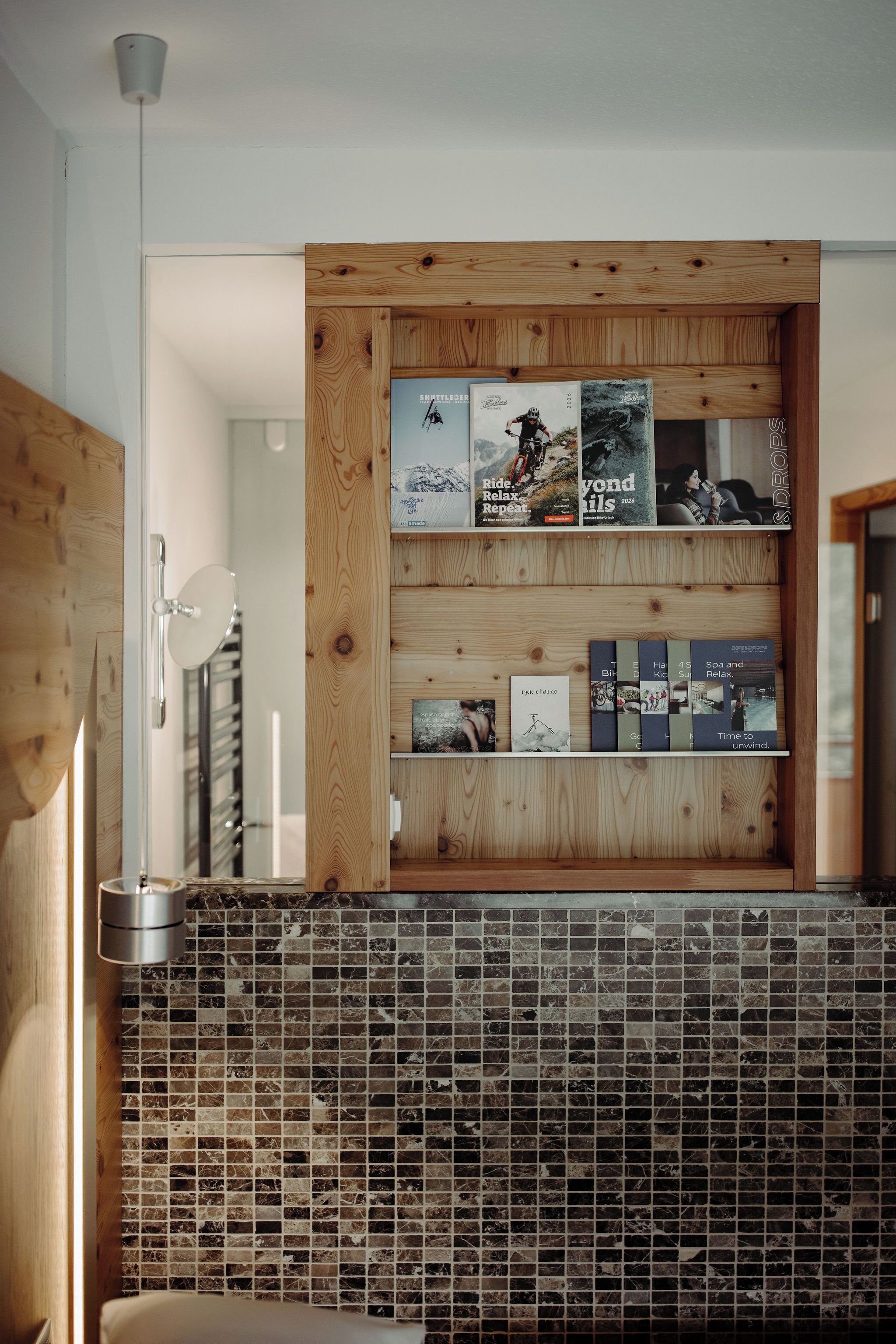 A modern bathroom with a wooden vanity, a tiled backsplash, and a shelf with magazines.