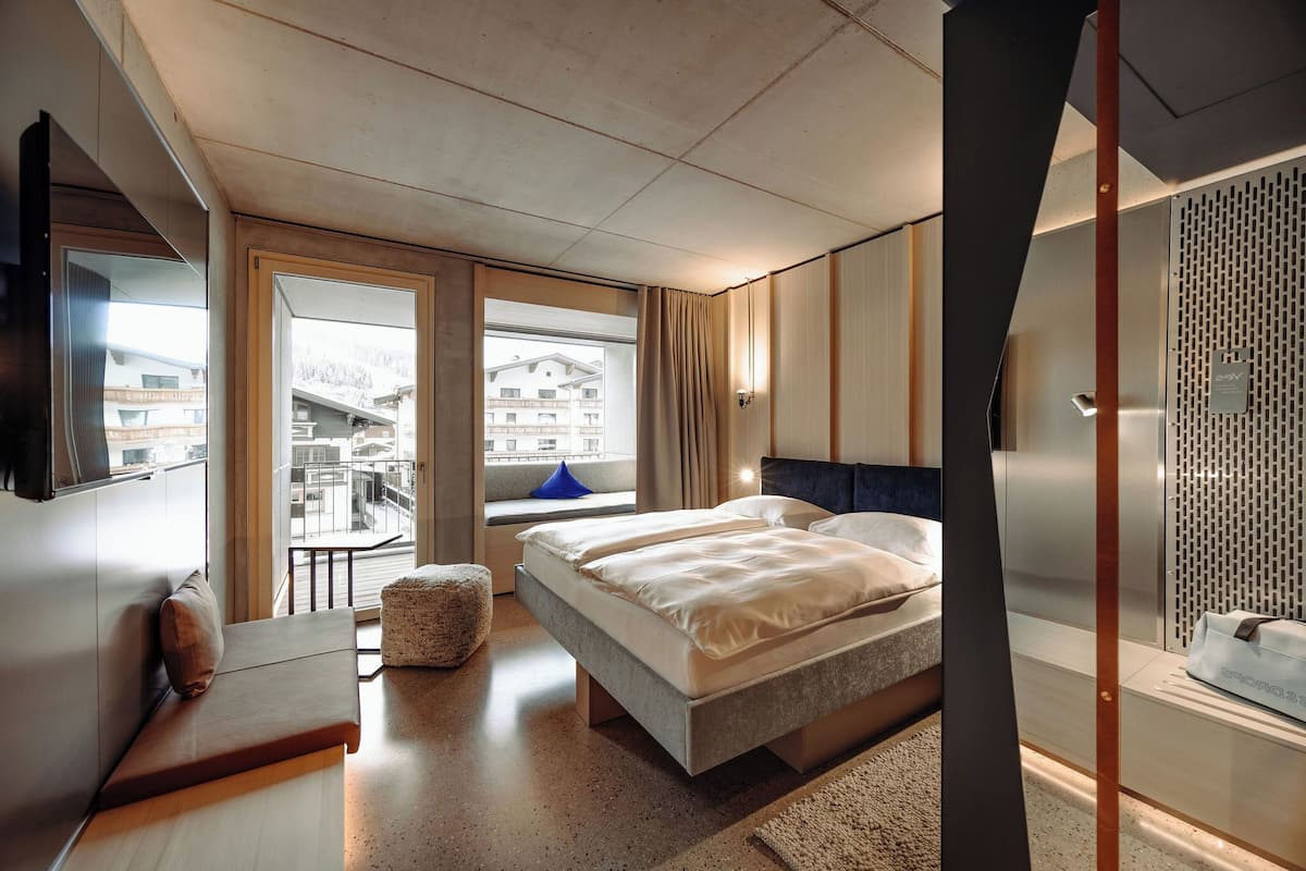 A modern hotel room with a large bed, a seating area, a TV, and a view of the city.