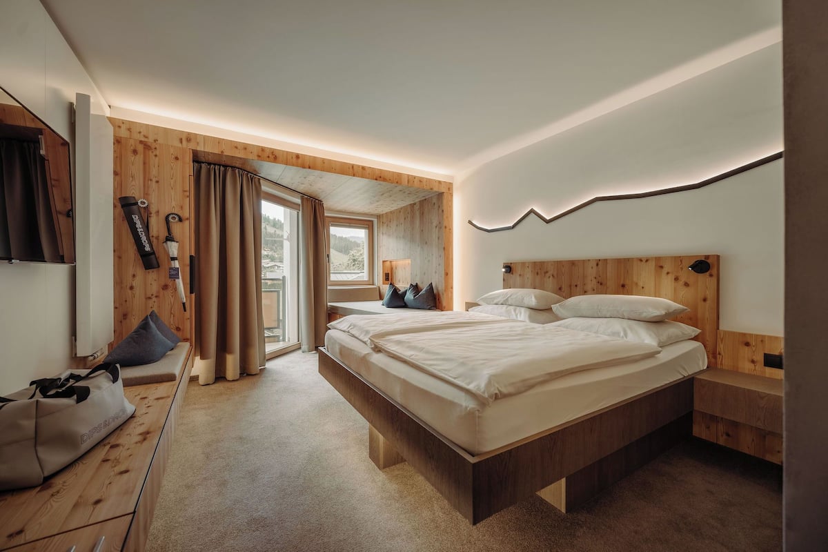 A bedroom with a wooden headboard, a bed with multiple pillows, a bedside table, a window with curtains, and a view of the outdoors.