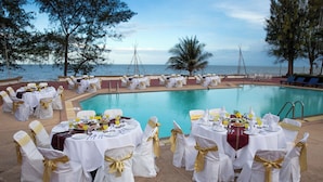 Zona de comedor junto a la piscina con mesas preparadas para una comida, con vistas a una masa de agua.