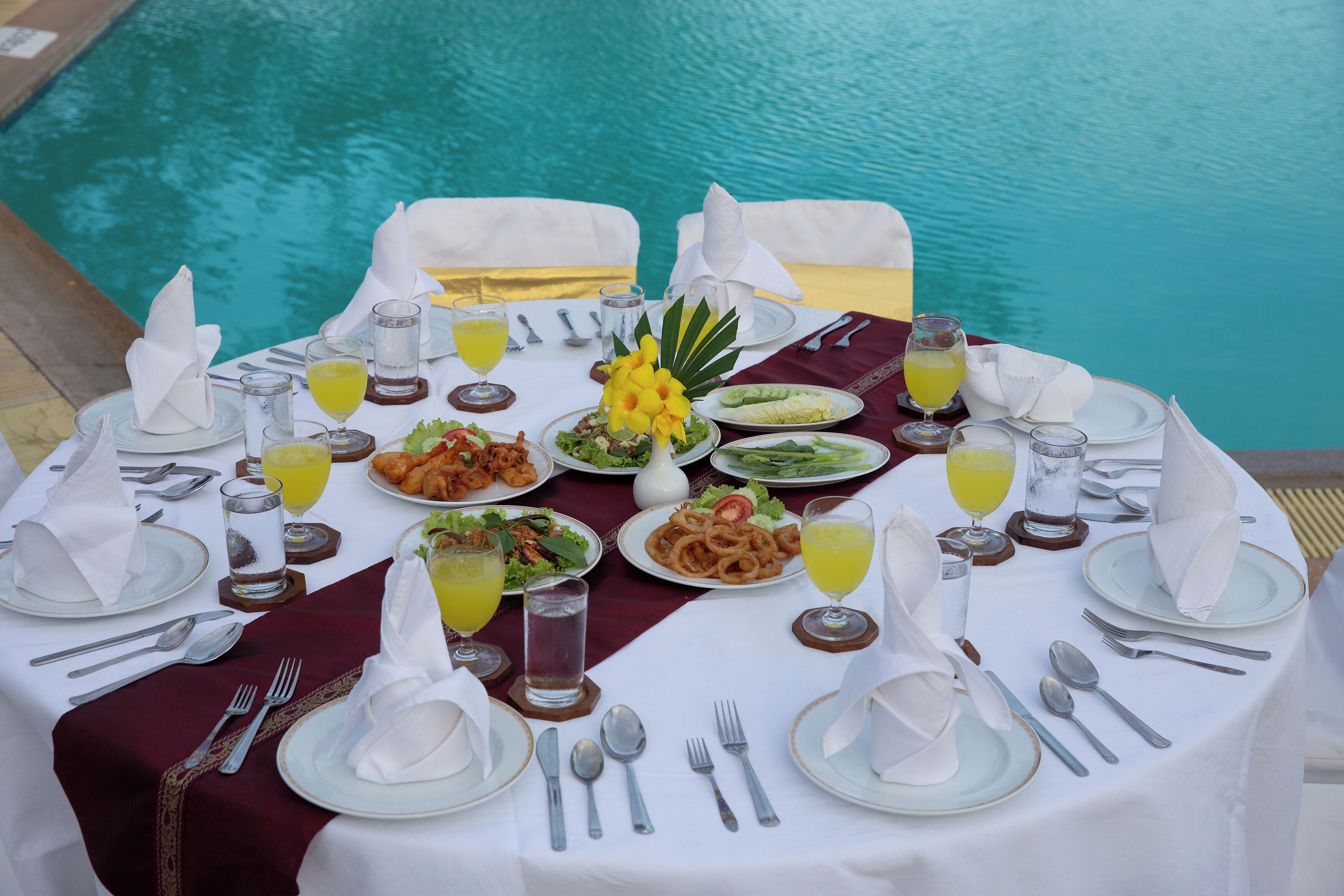 A table set for a meal by a poolside with various dishes and drinks.