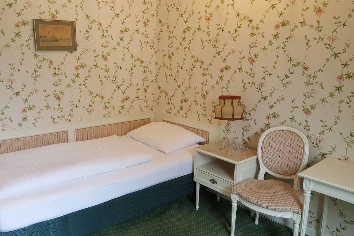 Une chambre avec un lit simple, une chaise, une table de chevet et une lampe fixée au mur.