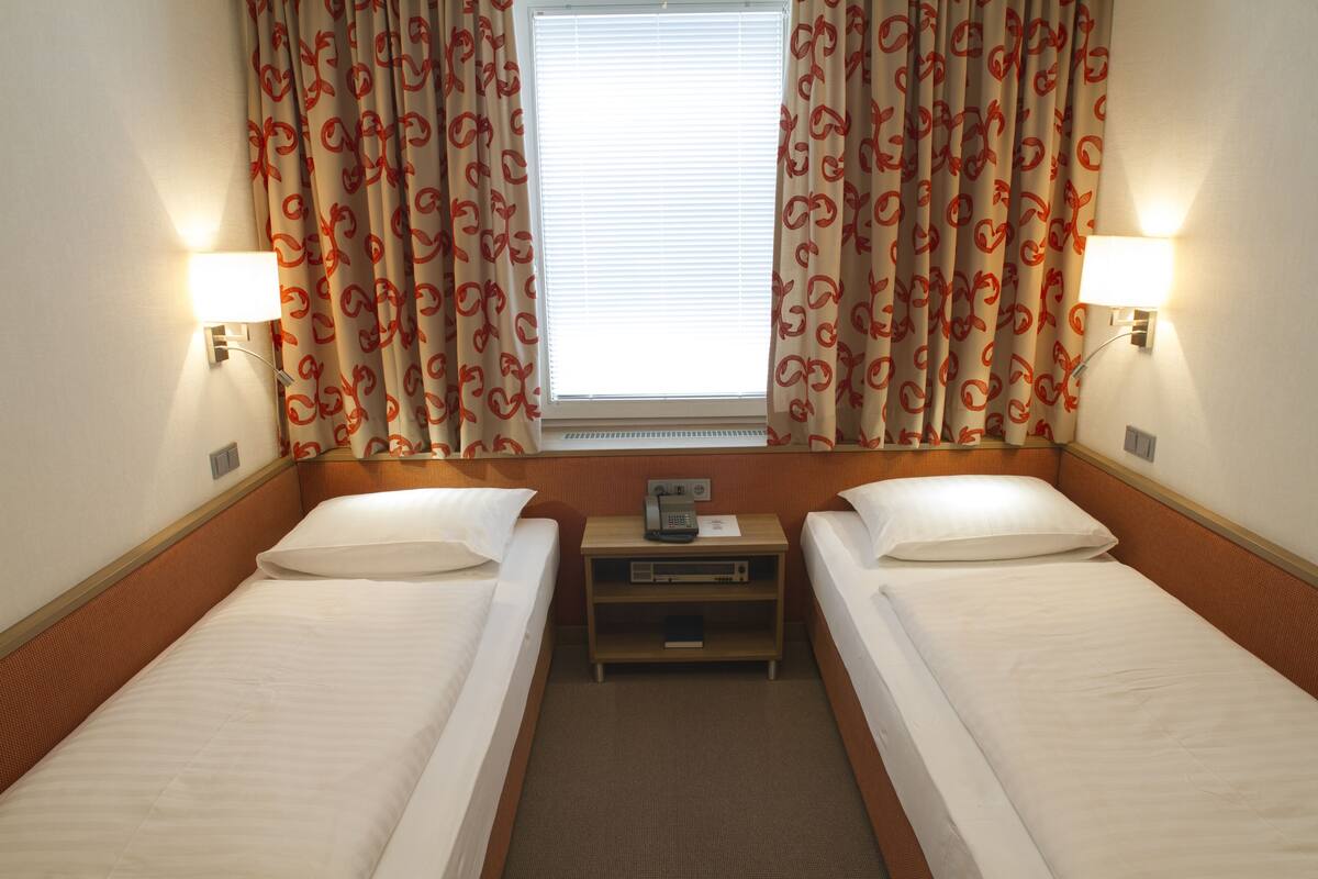 Une chambre d’hôtel avec deux lits simples, une table de chevet avec une lampe, un téléphone et une petite étagère.