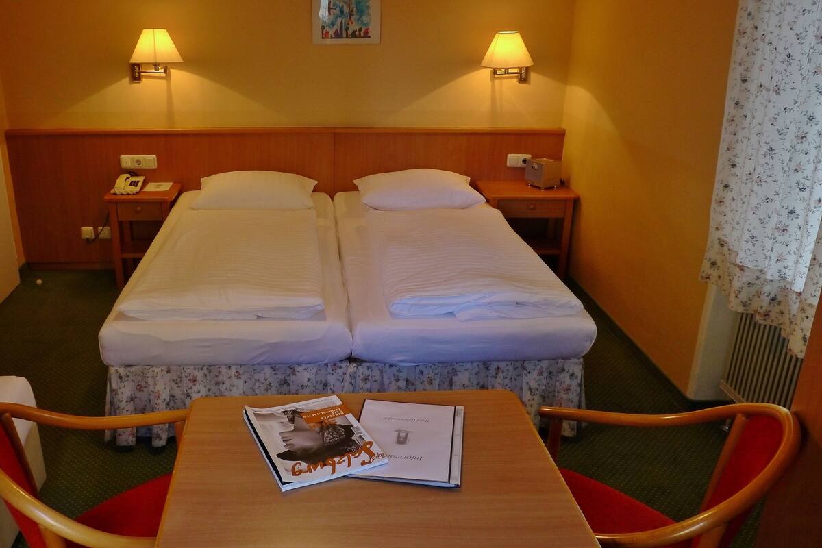 Une chambre d’hôtel avec deux lits, une table avec un magazine, deux tables de chevet avec des lampes et une porte.
