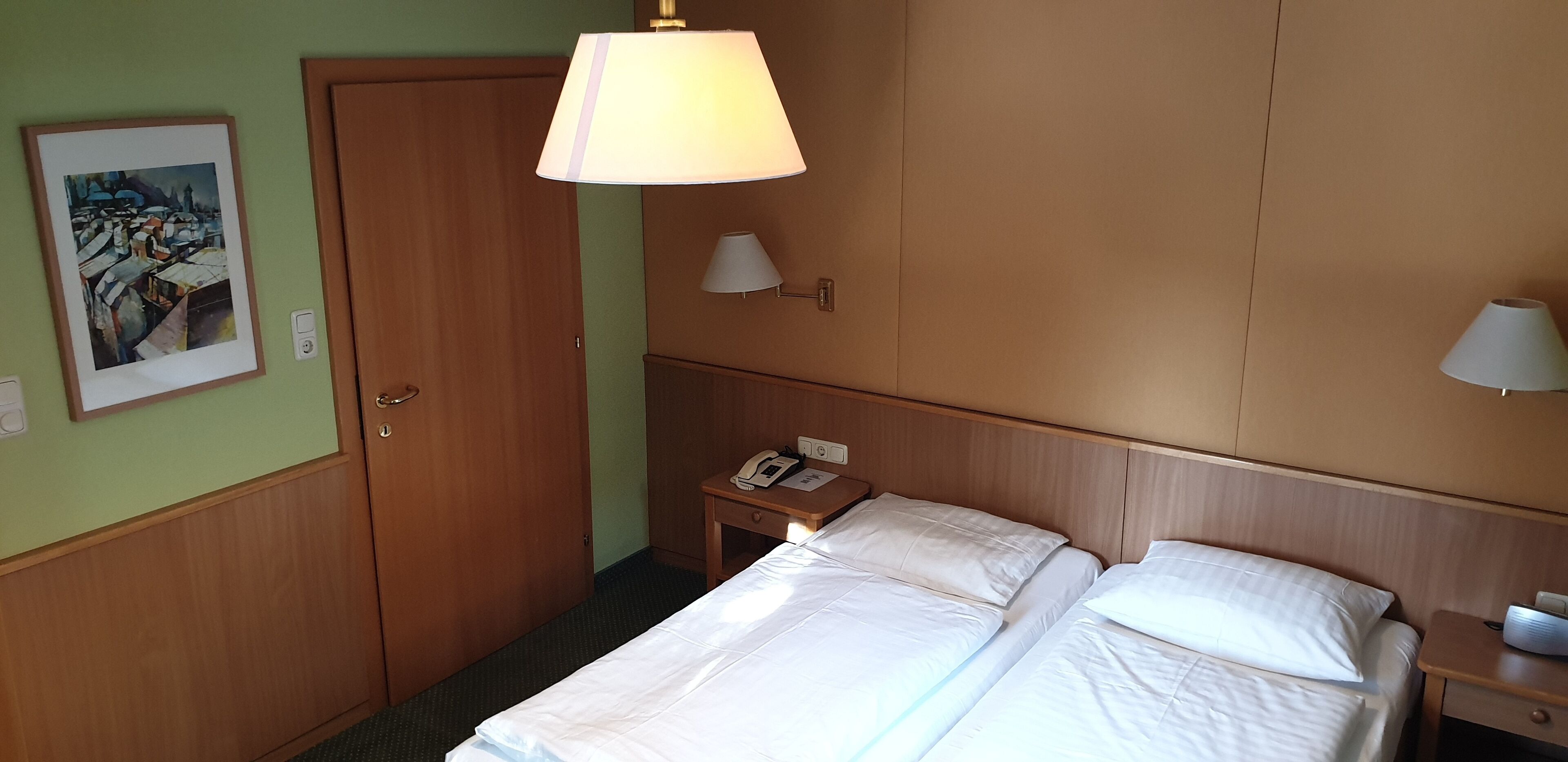 Una habitación de hotel con cama, mesitas de noche, lámpara, un cuadro en la pared y una puerta.