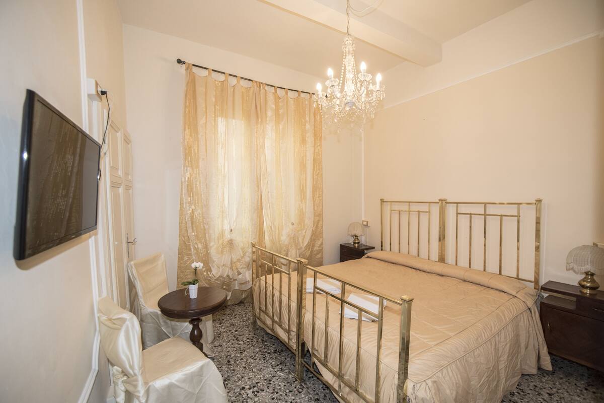 A bedroom with a bed, a chandelier, a television, a chair, a table, and a curtain.