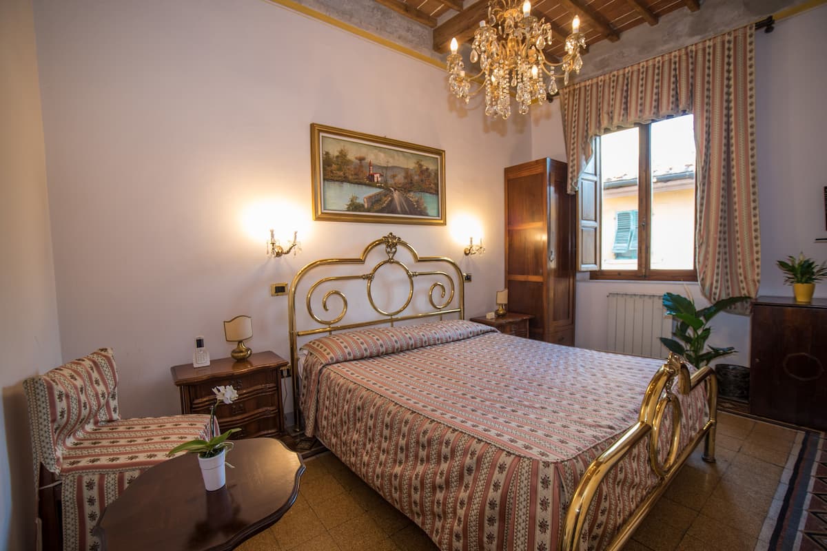 A bedroom with a bed, a chair, a nightstand, a dresser, a chandelier, a window with curtains, and a painting on the wall.