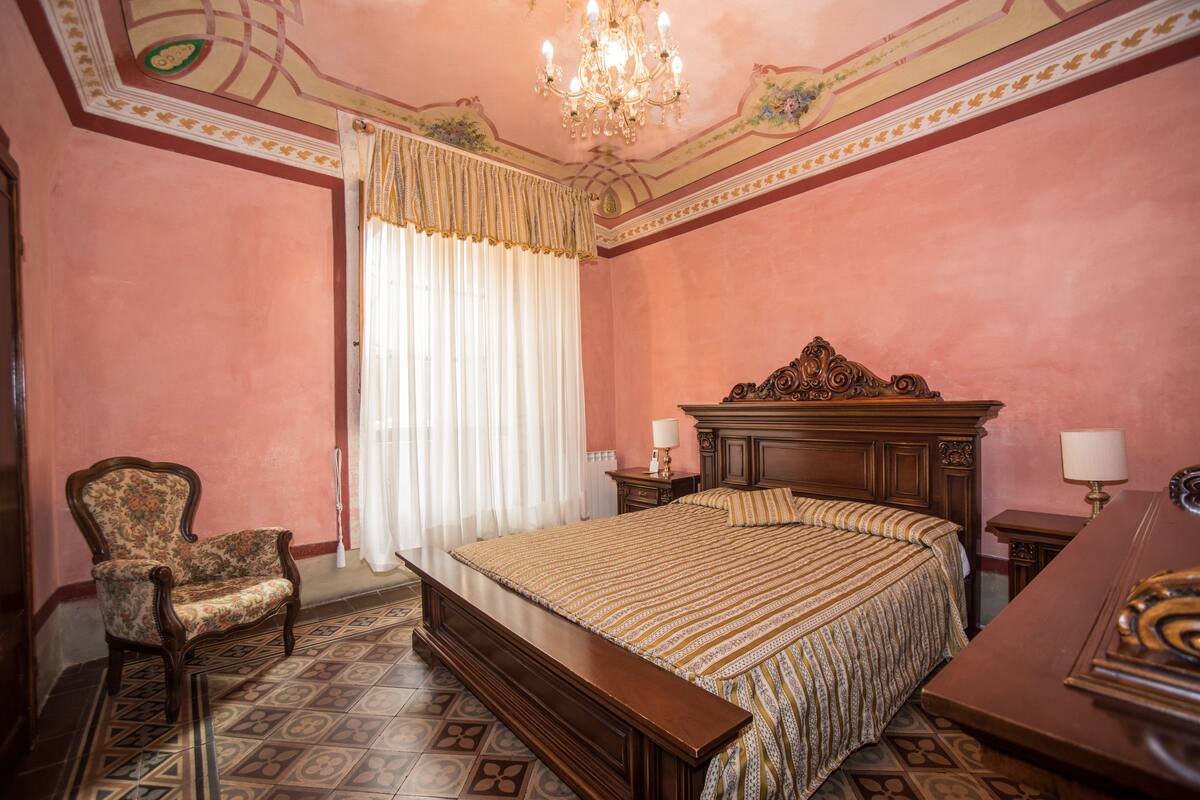 A bedroom with a large bed, a wooden headboard, a chair, a nightstand, and a chandelier.
