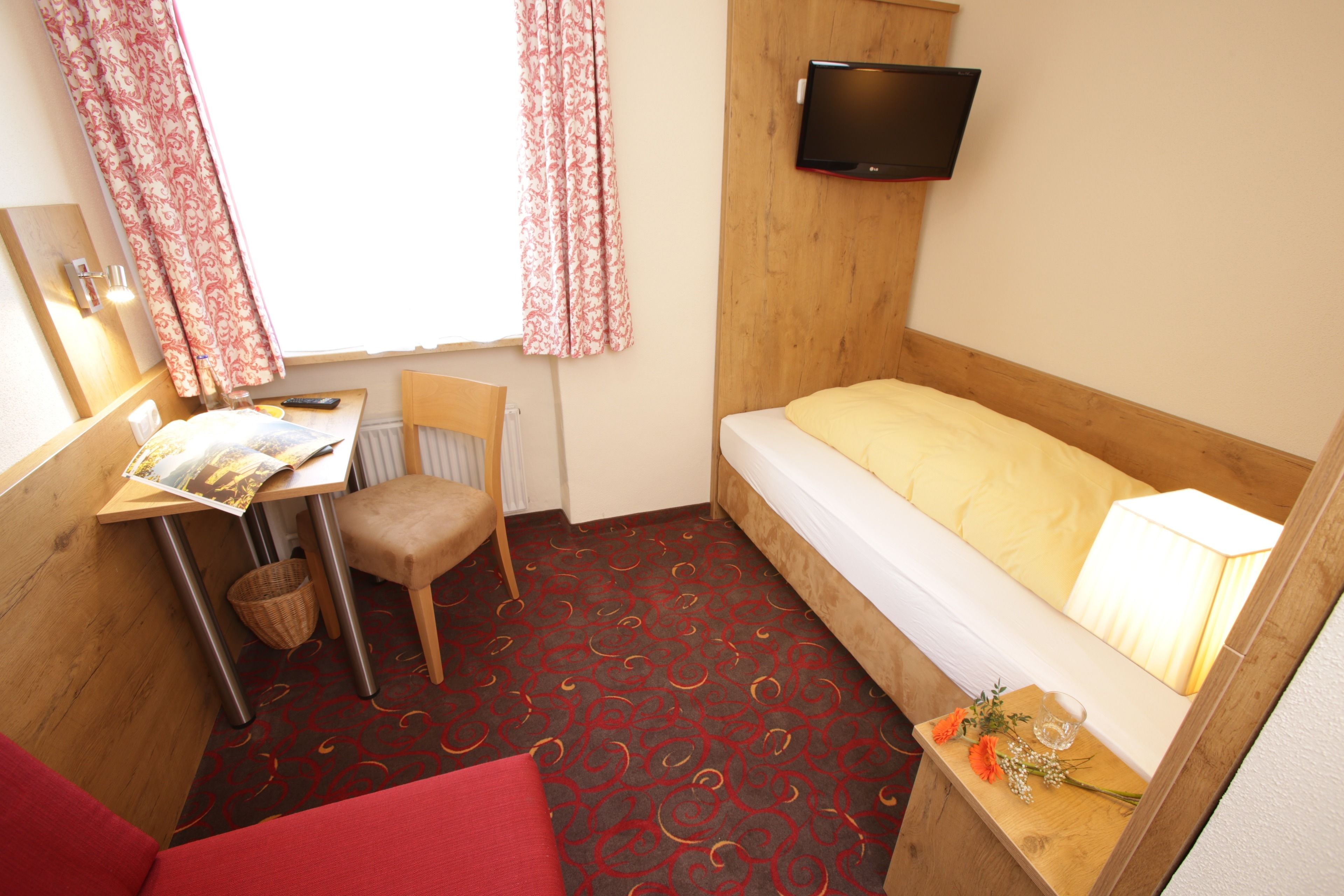 A small, compact room with a single bed, a desk with a chair, a television mounted on the wall, and a window with curtains.