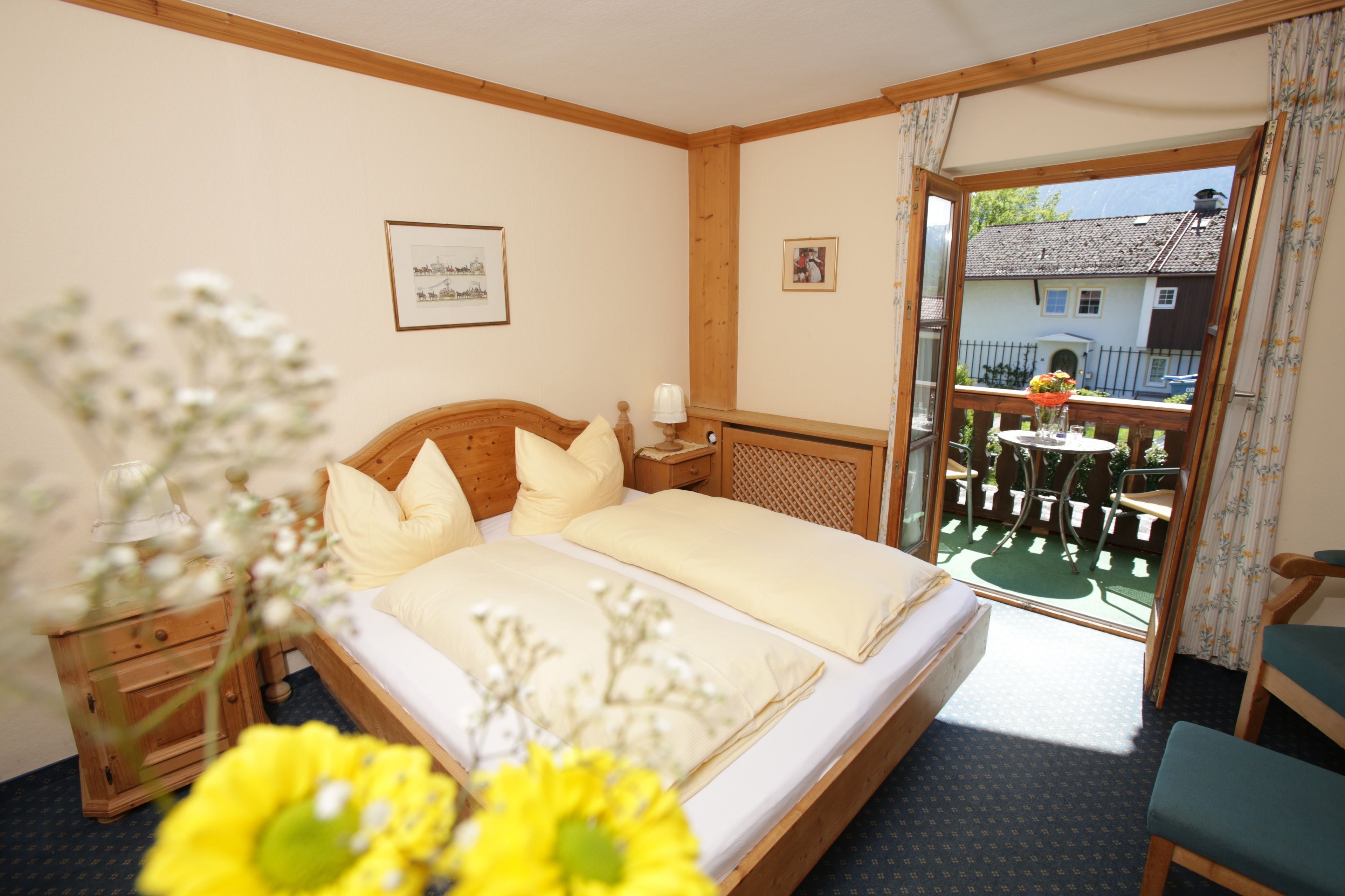 A bedroom with a wooden bed, a nightstand, a window with a view of a house, and a balcony with a table and chairs.