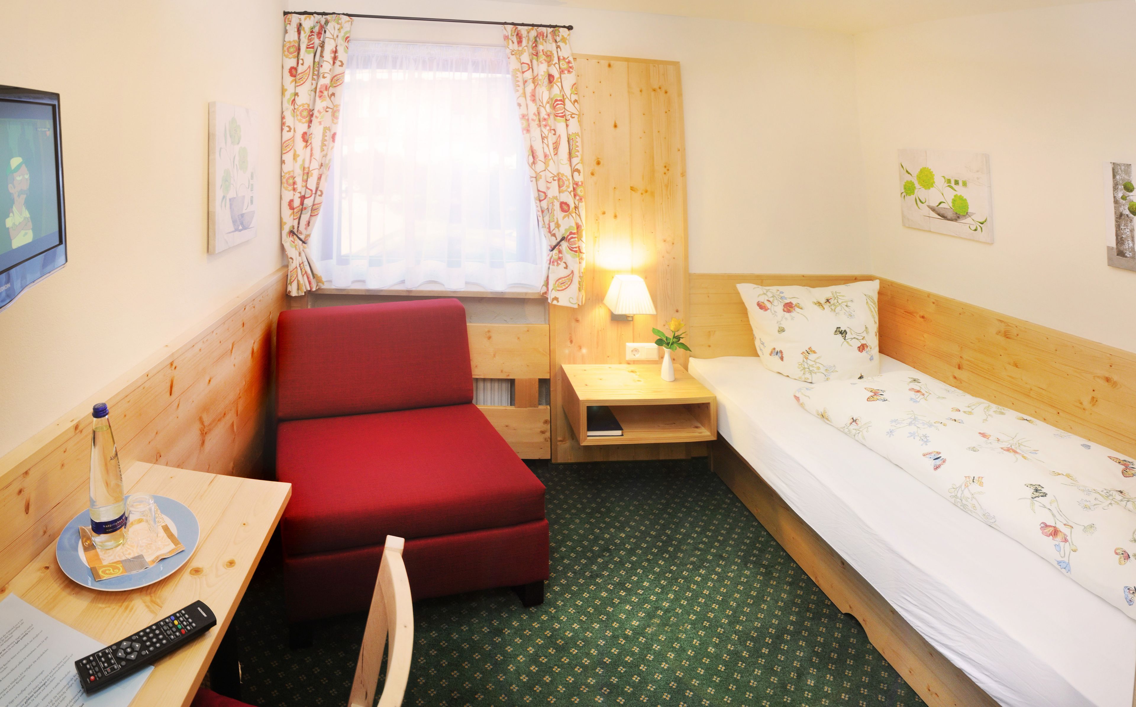 Una habitación de hotel pequeña con una cama individual, una silla roja, una mesita de noche de madera con una lámpara, una pequeña mesa con un control remoto y una ventana con cortinas florales.