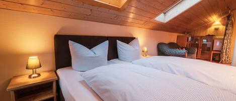 Un dormitorio con techo de madera, tragaluz, cama con ropa de cama a rayas, mesita de noche con lámpara y sillón con cojín.