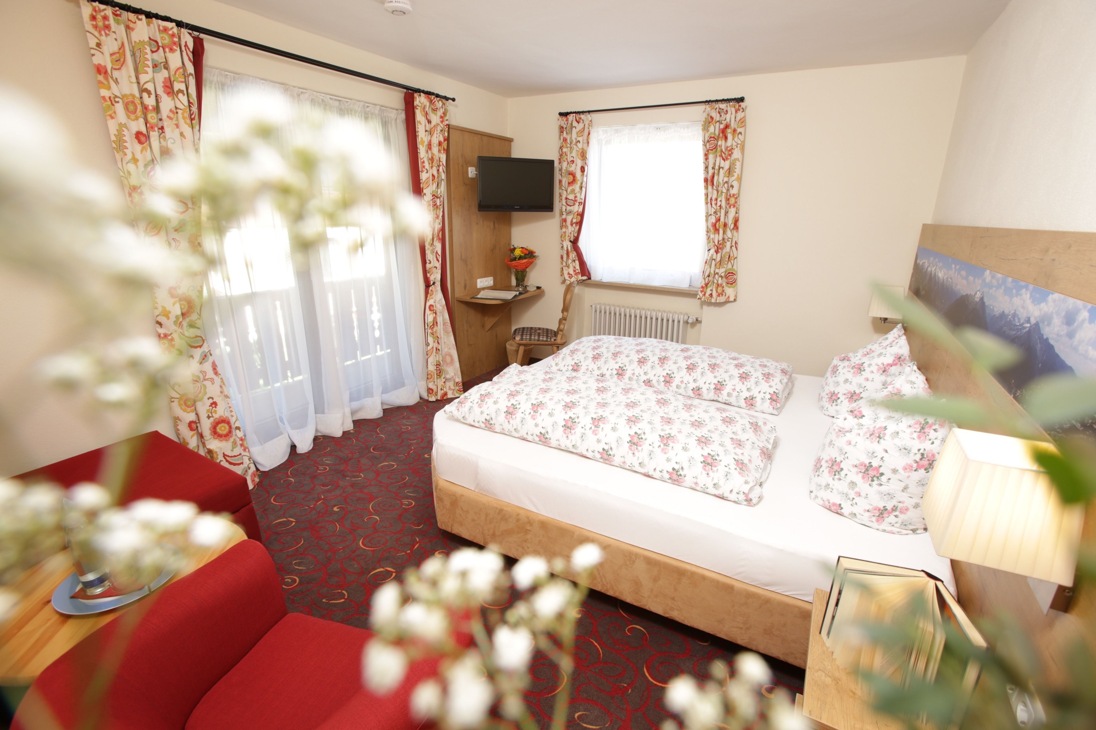 Una camera d'albergo con un letto, una televisione fissata al muro, una finestra con tende e un divano rosso.