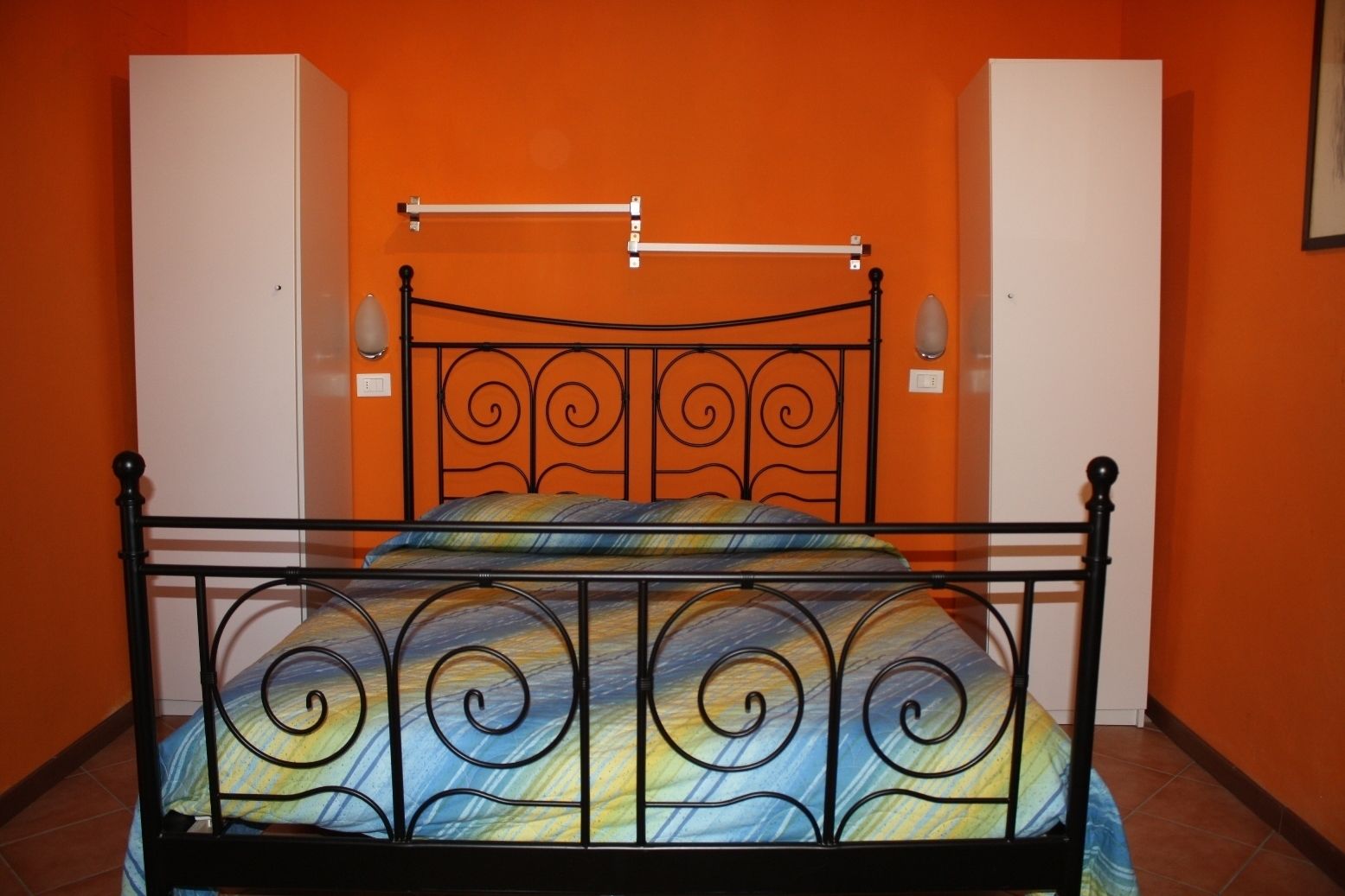 A single bed with a metal frame and a colorful bedspread in a room with orange walls and white cabinets.