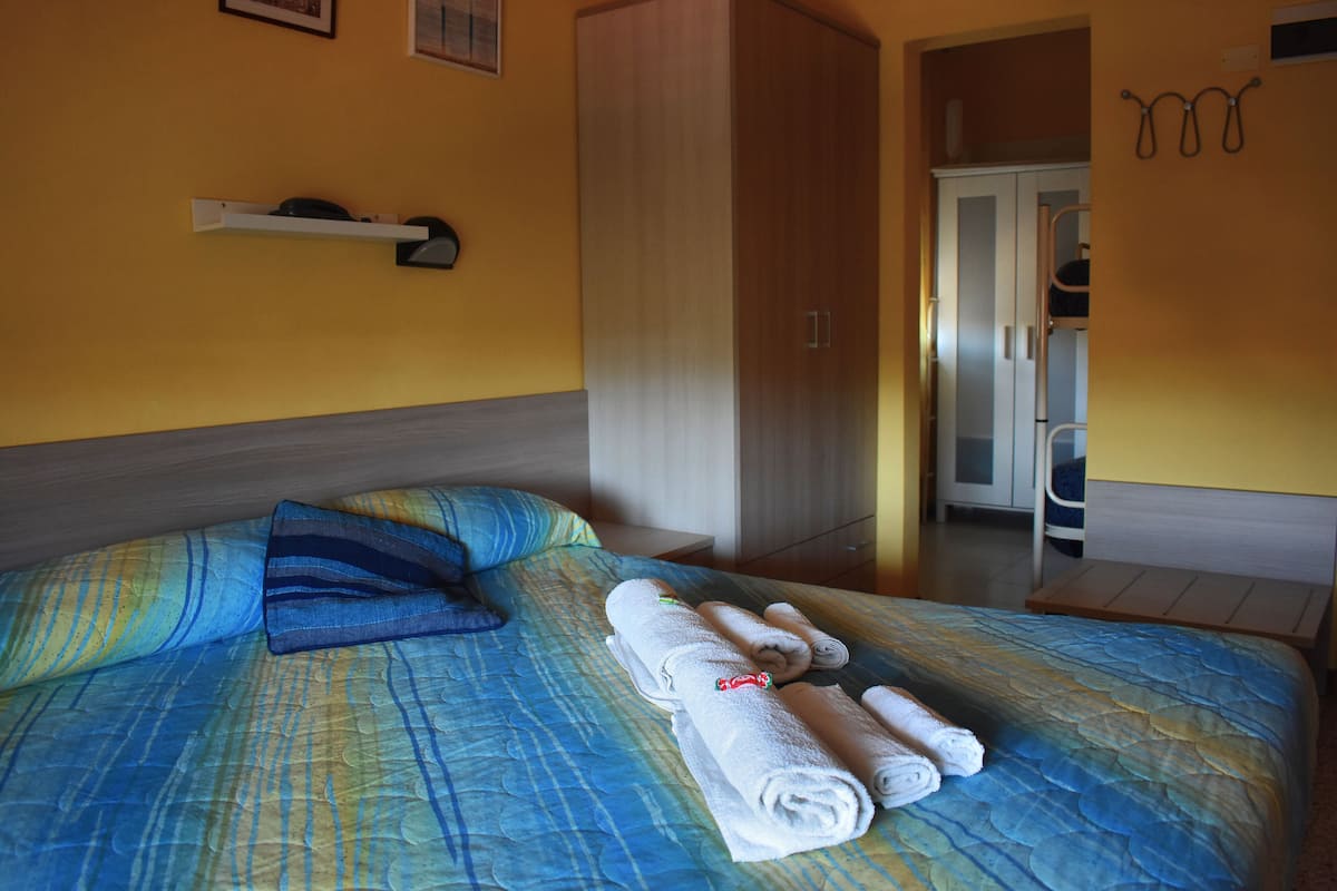 A neatly made bed with blue and yellow bedding, a wooden wardrobe, and a small white shelf against a yellow wall.