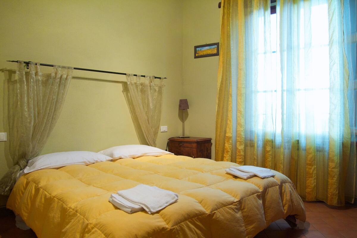 A bed with a yellow bedspread and white pillows, a small wooden nightstand with a lamp, and sheer curtains allowing natural light.