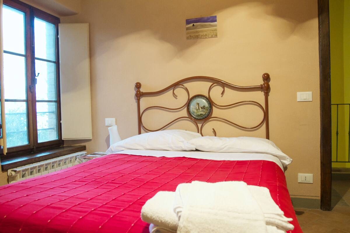 A bedroom with a red bedspread, a wooden headboard with a decorative plate, white pillows, and a window with blinds.
