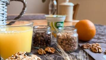 A breakfast setting with a glass of orange juice, a loaf of seeded bread, a jar of nuts, a whole orange, and a vase with dried wheat stalks.