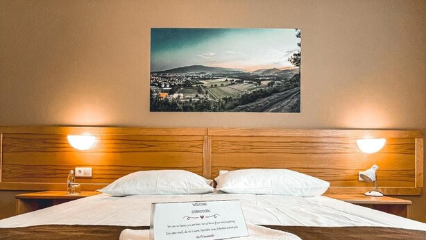 A neatly made bed with a welcome sign, a framed landscape photo on the wall, and two bedside lamps. - Hotel Bau (Maribor)