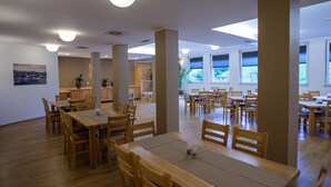 A spacious dining area with wooden tables and chairs, large windows, and a view of the outdoors. - Hotel Bau (Maribor)