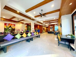 A spacious lobby with high ceilings, wooden beams, and ceiling fans. Benches with colorful cushions line the walls, and a reception desk is visible in the background. - Baramee Resortel (Patong)