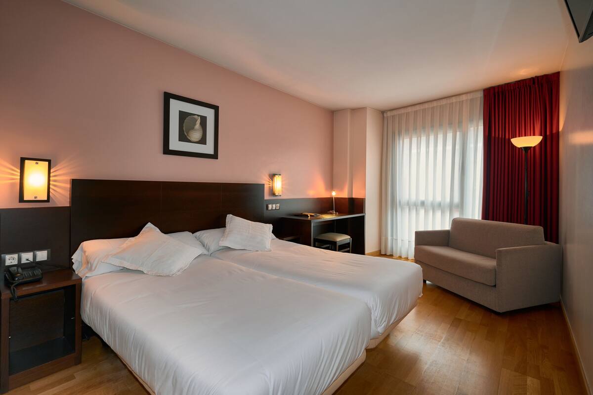 A hotel room with a large bed, two bedside tables, a desk, a chair, and a window with curtains.