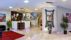 Un vestíbulo de hotel con mostrador de recepción, una escalera, un espejo grande y un área de estar con alfombra roja.