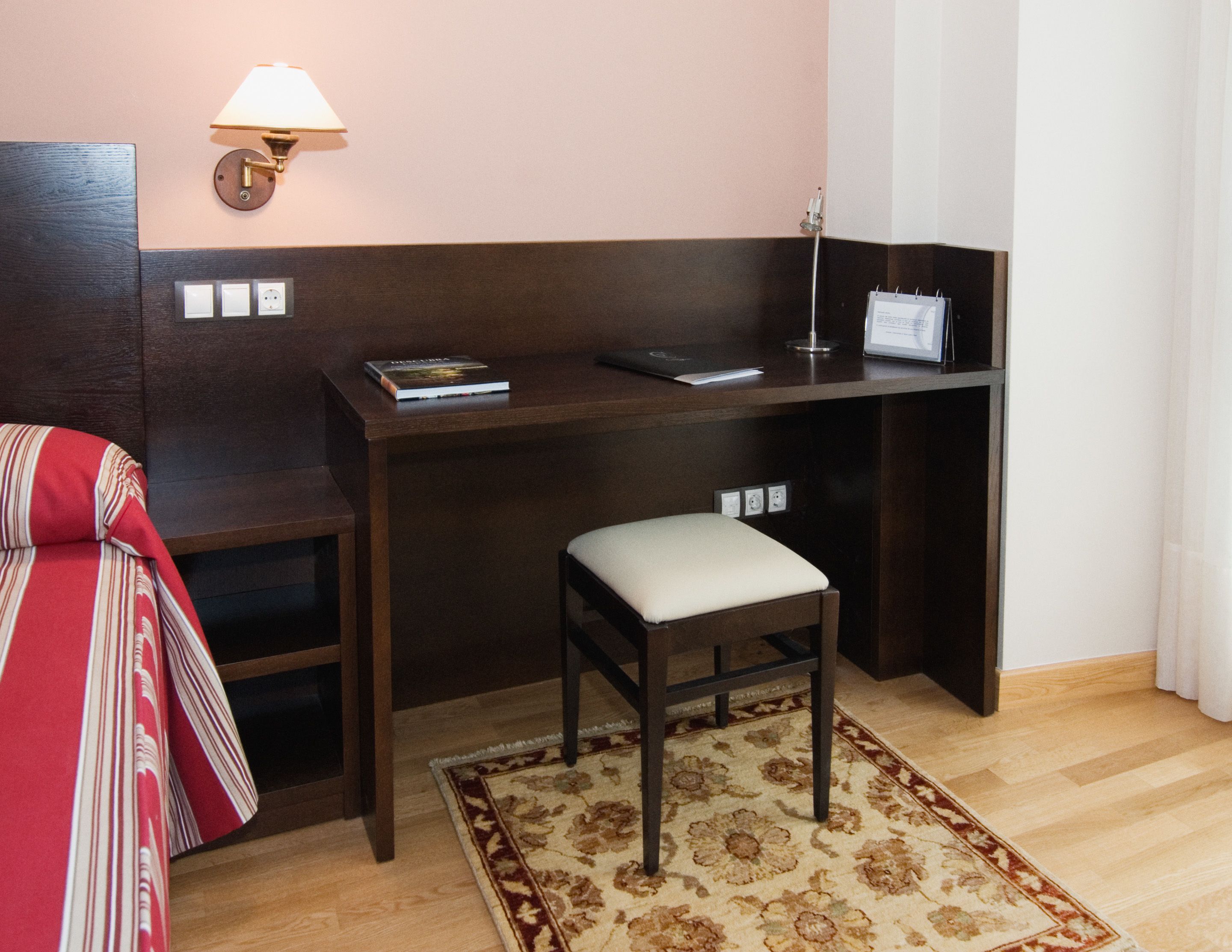 Una habitación de hotel con un escritorio de madera, un taburete pequeño, una lámpara y un sofá a rayas rojas.
