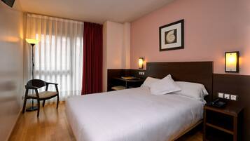 A hotel room with a large bed, a chair, a desk with a lamp, and a nightstand with a phone.