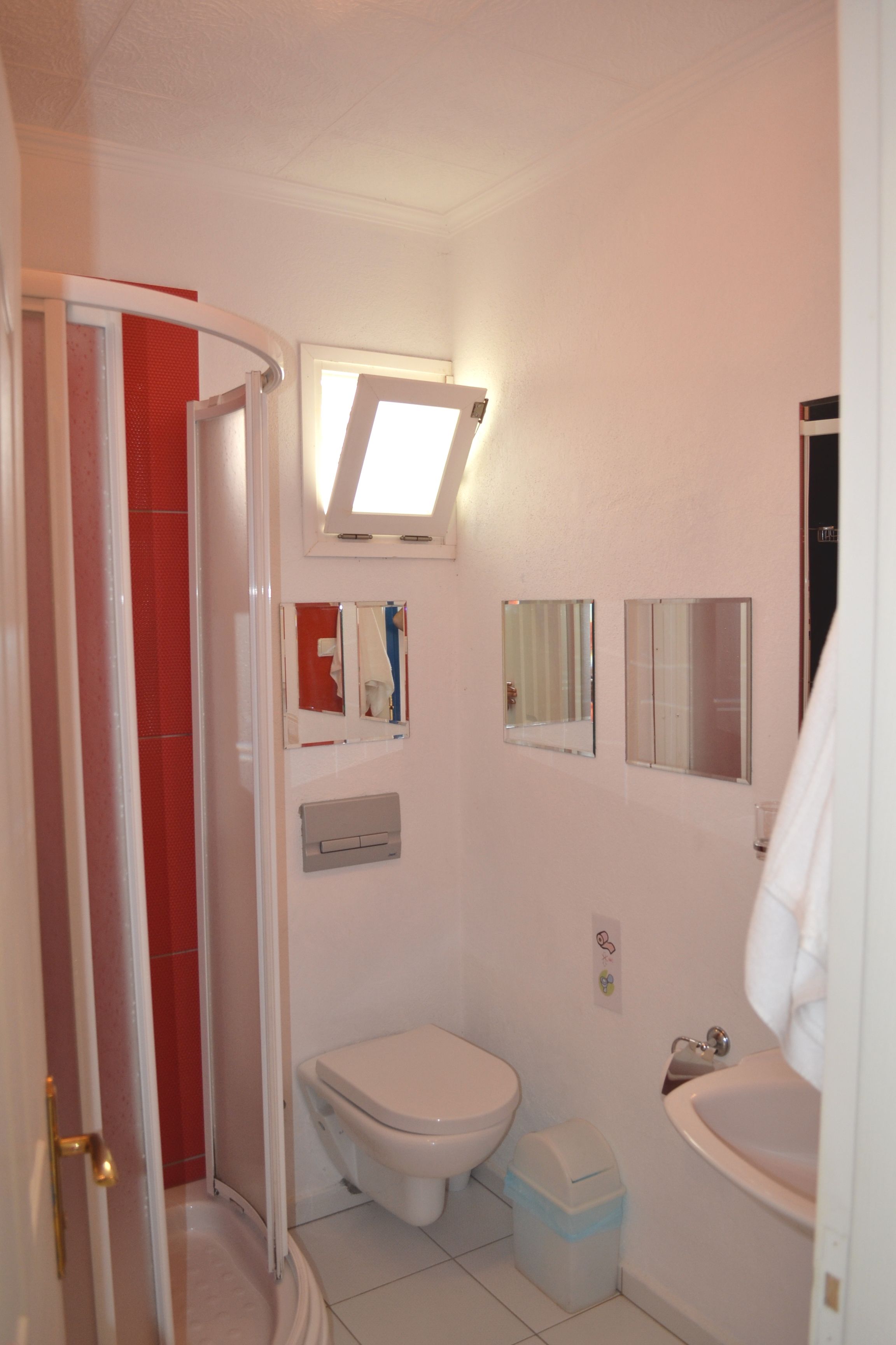 A bathroom with a toilet, sink, and shower. There are two mirrors on the wall and a window above the toilet.