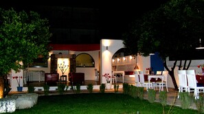 An outdoor dining area with tables and chairs, a stone wall, and a well-lit entrance.