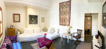 Una sala de estar con un sofá blanco, una mesita con una lámpara, una silla y un tapete grande. Hay cuadros enmarcados en las paredes y una puerta que da a otra habitación.