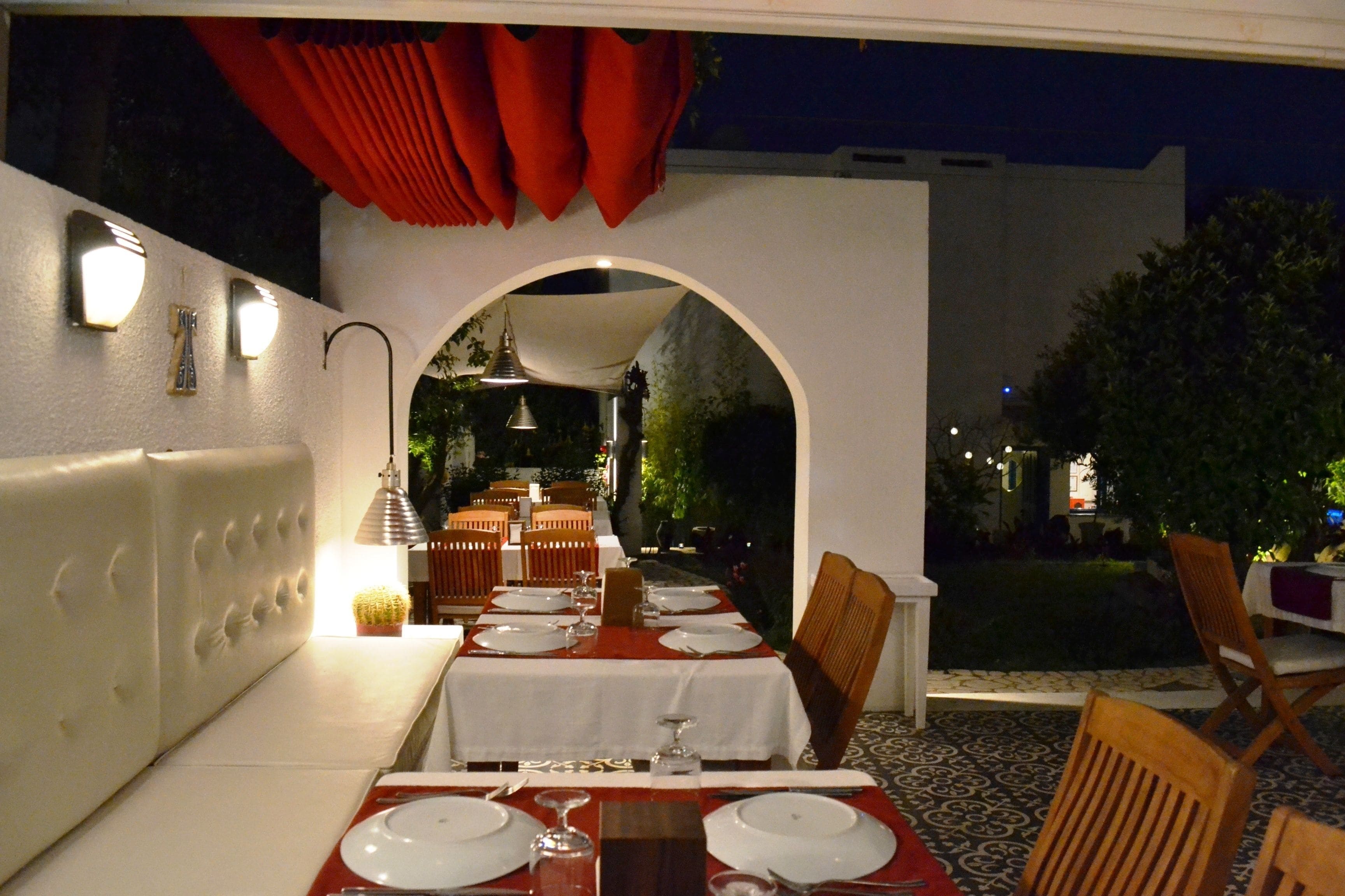 An outdoor dining area with tables set for a meal, a white cushioned bench, and a red curtain above the entrance.