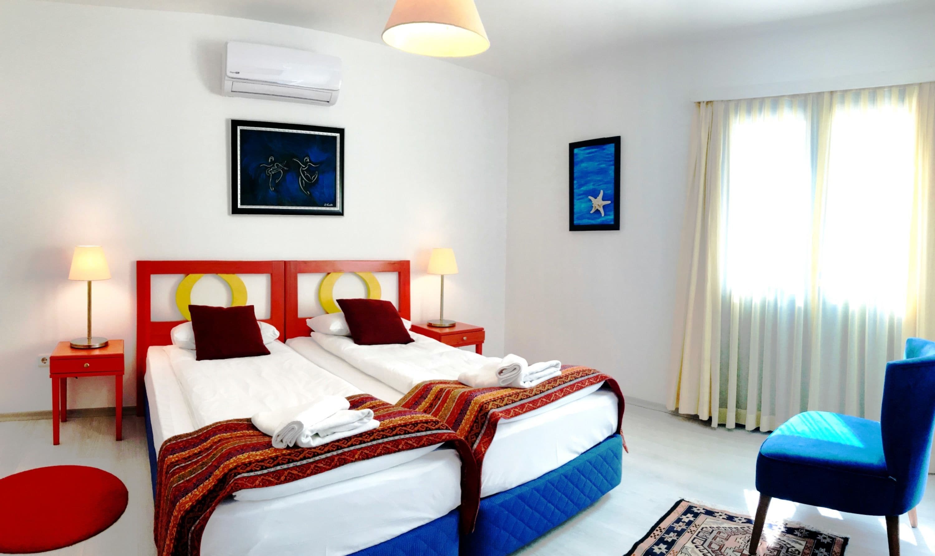 Habitación de hotel con una cama doble, dos mesitas de noche con lámparas, cabecera con marco rojo, una silla azul, una ventana con cortinas y dos cuadros en la pared.