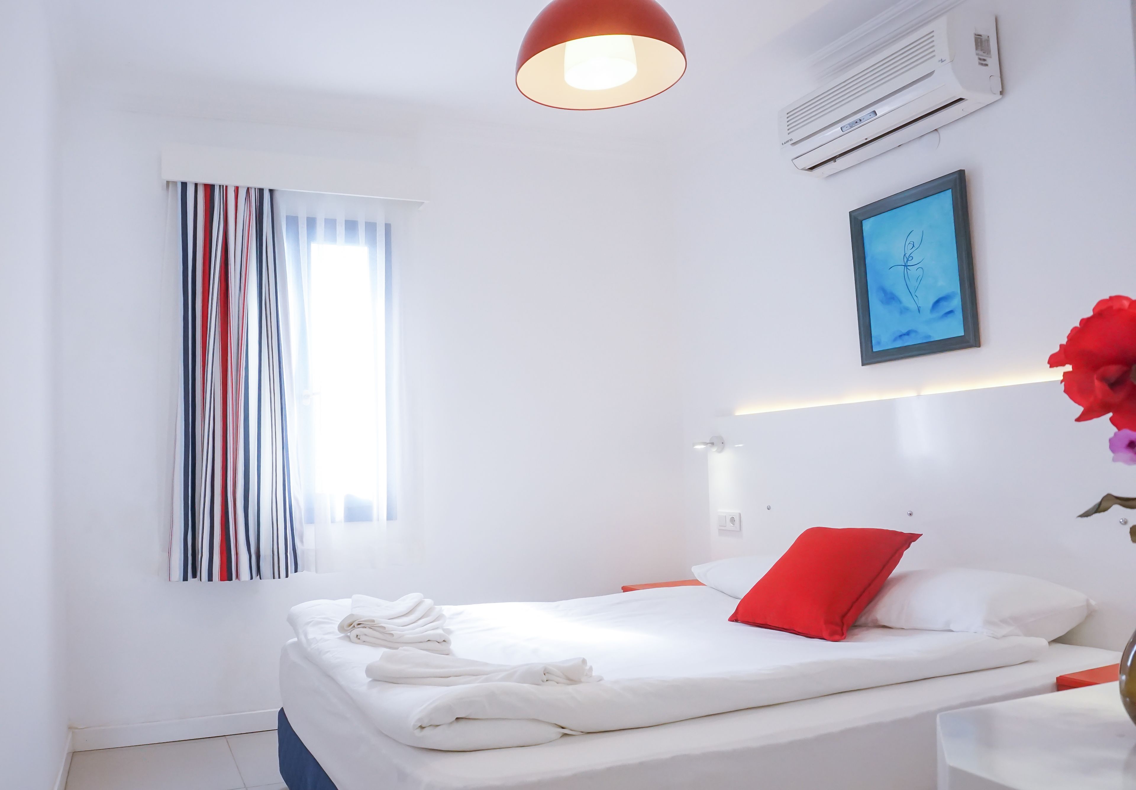 Una habitación de hotel moderna con una cama bien hecha, una almohada roja, una obra de arte enmarcada en la pared y una ventana con cortinas a rayas.
