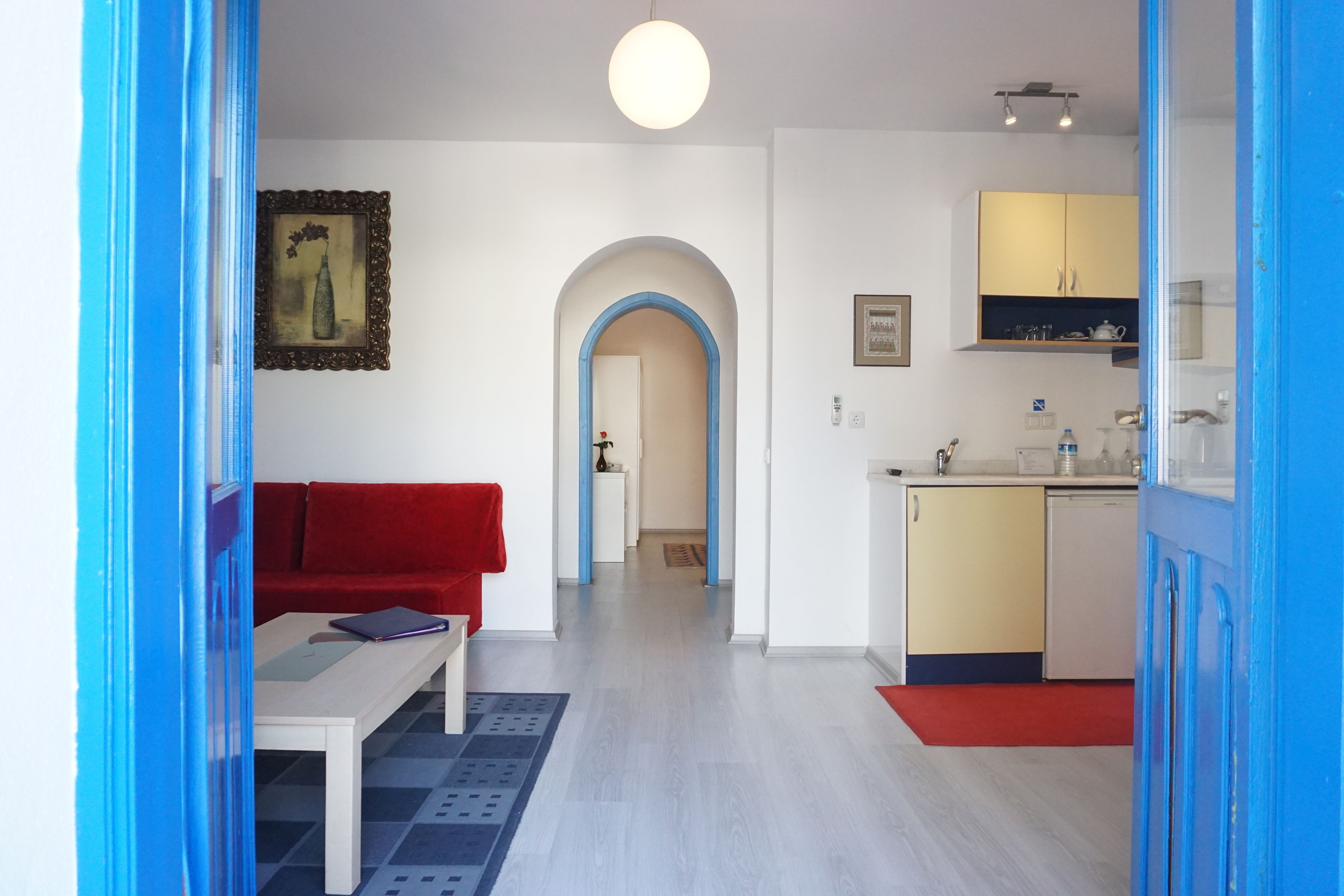 A modern living room with a red sofa, a white coffee table, and a blue patterned rug. There is a kitchen area with yellow cabinets and a white countertop.