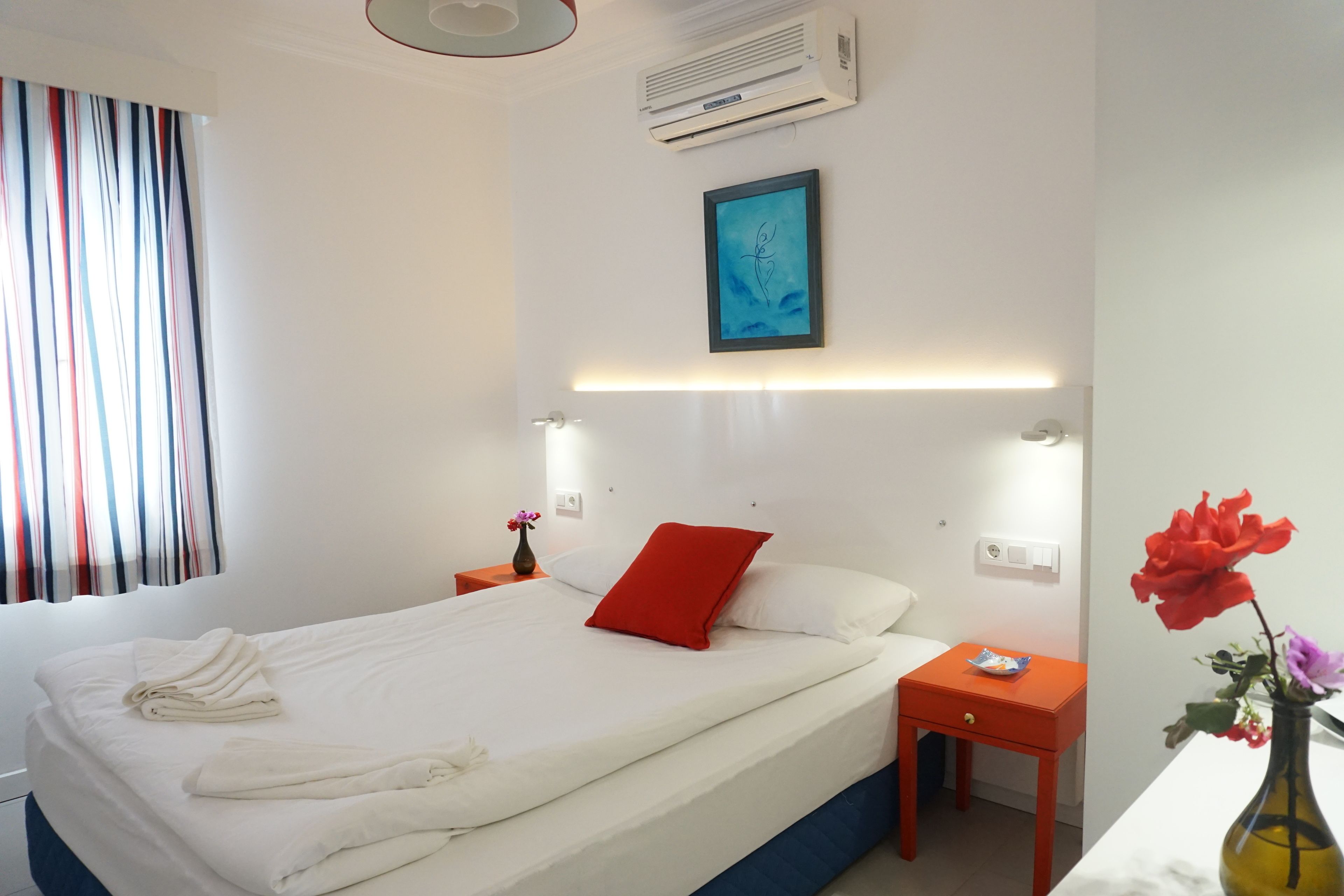 Una habitación de hotel con una cama bien hecha, una mesita de noche con un jarrón de flores y un aire acondicionado de pared.